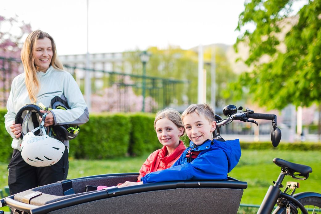kvinne står ved lastesykkelen sin der det sitter to barn oppi 